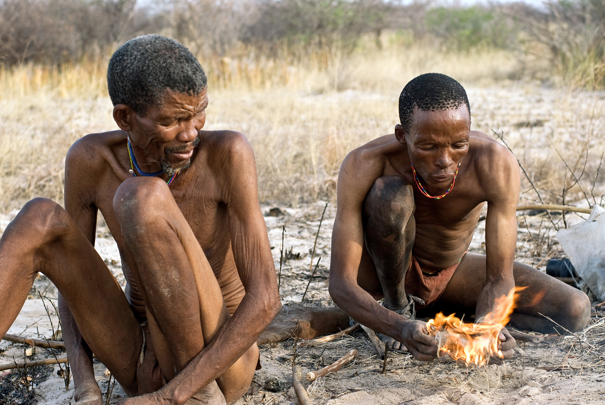 Fotografija prikazuje Bušmane, narod iz Afrike kako raspiruju vatru, Namibija. Dvojica muškaraca sjede na tlu i pokušavaju raspiriti vatru.